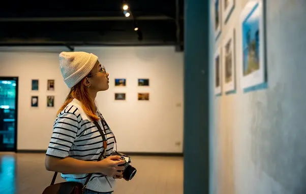 Asian woman hold camera at art gallery collection in front framed paintings pictures on wall